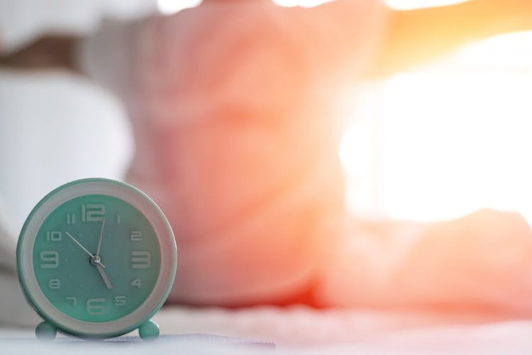 An image of a teal colored clock set to 5 am with a person waking up and performing morning stretches in the background. 