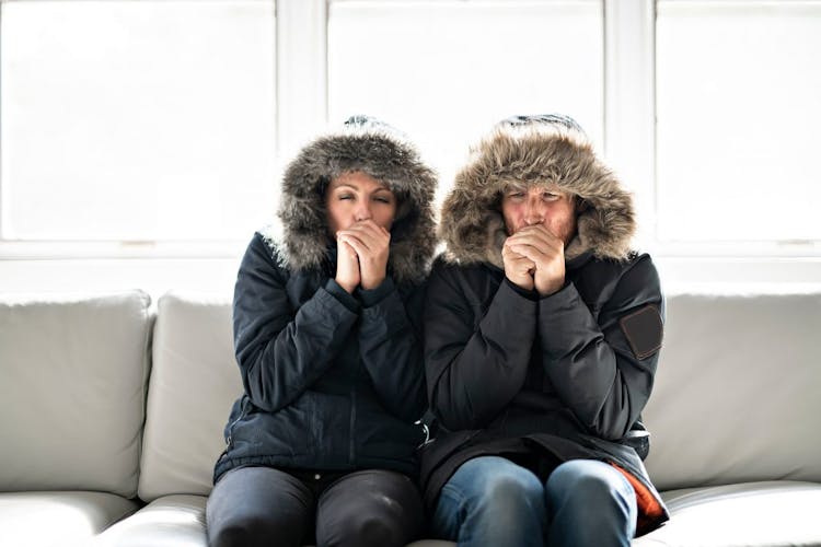 A man and a woman sitting on a couch in winter coats with their hoods up, blowing warm air into their cold hands