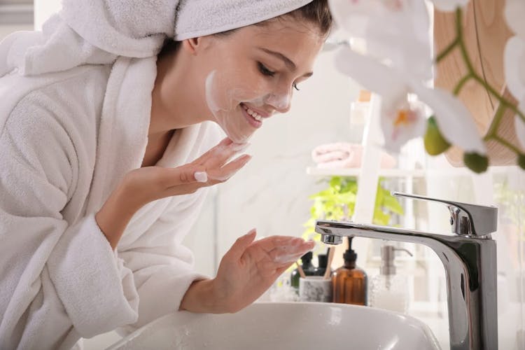 A smiling young woman standing over the sink in her bathroom cleansing her face using a gentle cleanser 