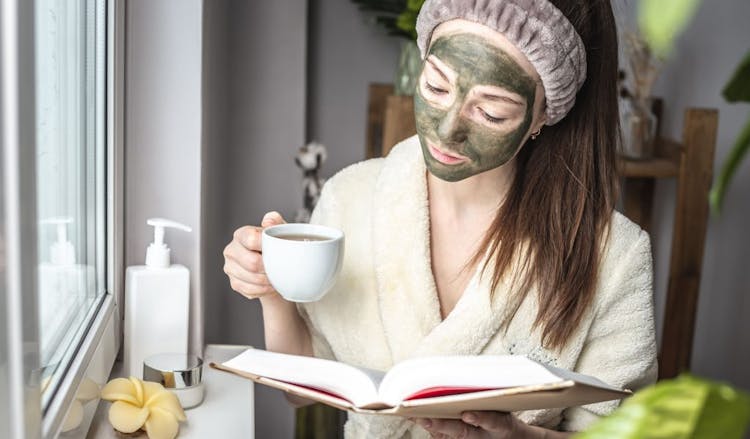 A young woman wearing a green tea face mask holding tea and reading a book with a hair net on 