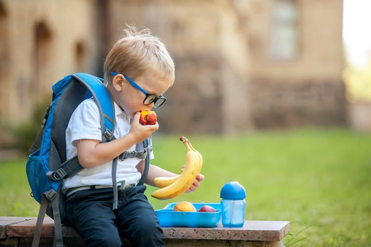 A cute young boy with a backpack on holding a banana and eating an apple at school during lunch