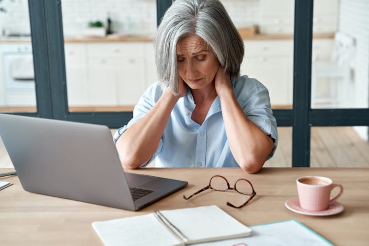 A senior woman working at her desk holding the back of her neck in pain