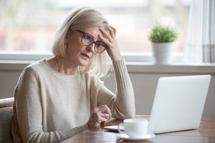 An older woman sitting at a table with her computer drinking coffee and looking frustrated or forgetful
