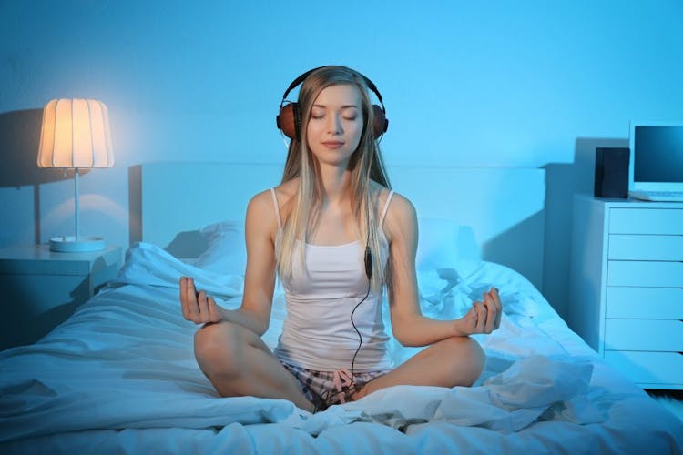 A young woman wearing headphones and meditating in bed 