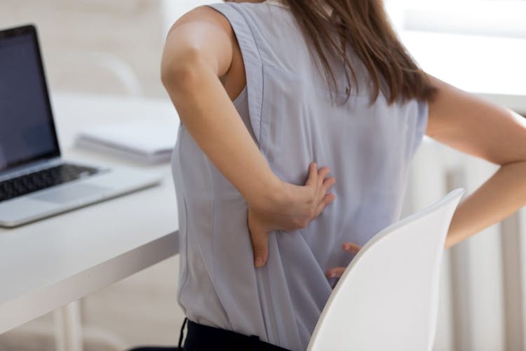 A woman sitting in a chair at her desk experiencing back pain and stiffness