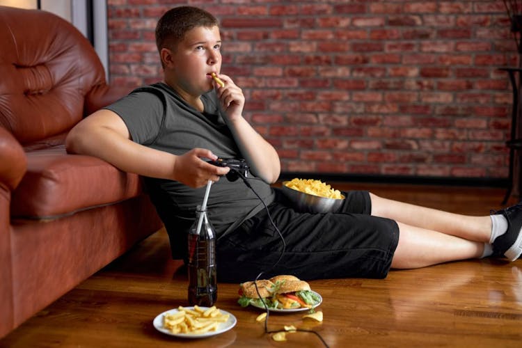 An overweight teenage boy eating unhealthy food and drinking soda while playing video games