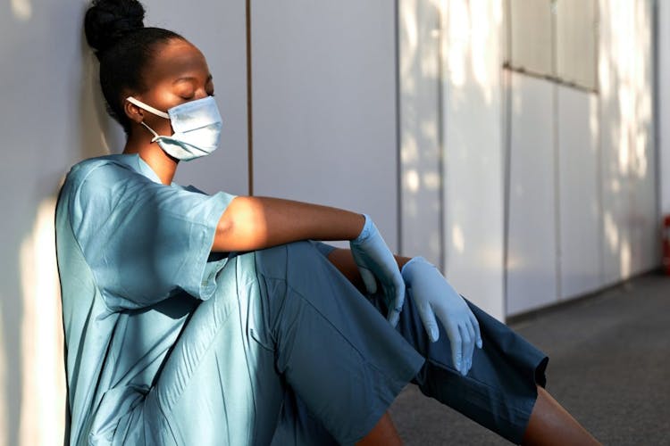 An exhausted nurse with a mask on sitting on the floor closing her eyes 