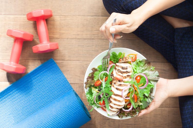 An image of a woman eating a healthy chicken salad next to a yoga mat and exercise equipment