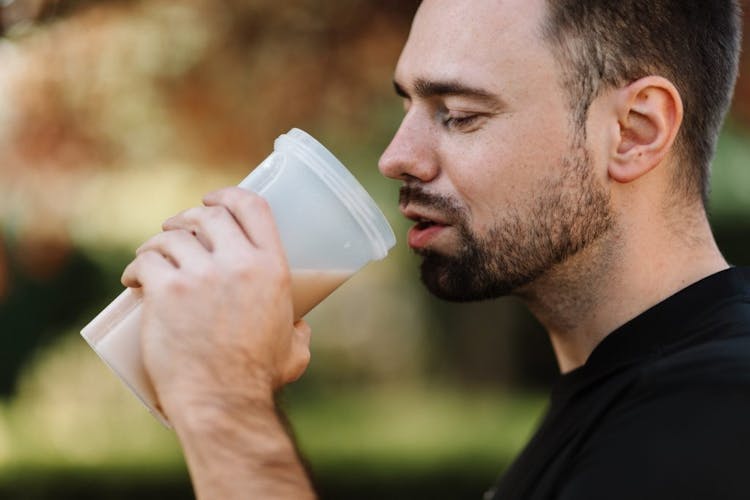 A man drinking protein drink from a transparent plastic tumbler.