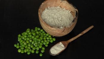 Pea protein powder is pictured in a wooden bowl, and in a wooden spoon from a side view. Next to them is a pile of peas.