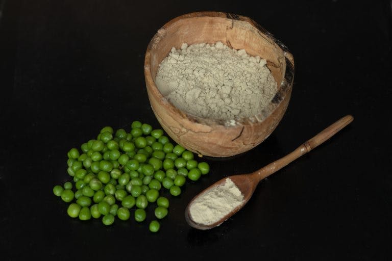Pea protein powder is pictured in a wooden bowl, and in a wooden spoon from a side view. Next to them is a pile of peas.