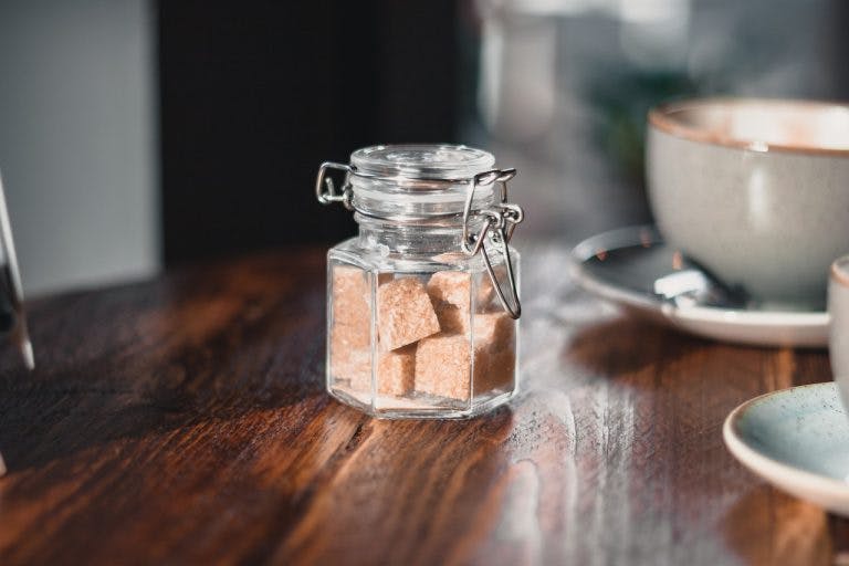 Clear condiment shaker with brown sugar cubes near gray teacup.