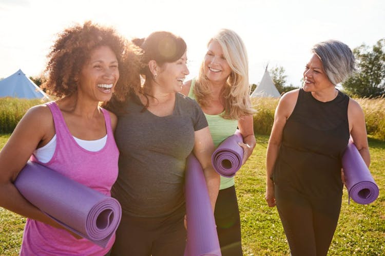 Four women wearing gym clothes laughing and talking outdoor, bringing yoga mat