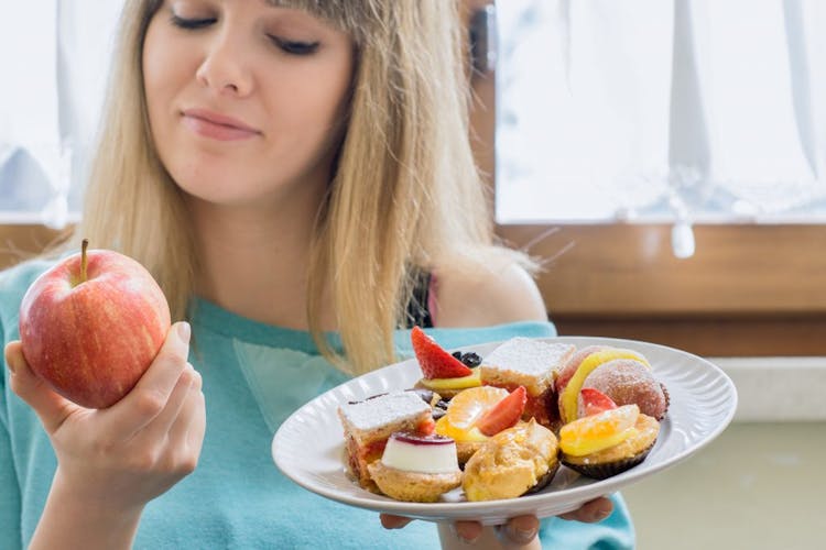 A woman holding an apple and a plate of desserts, contemplating a type 2 diabetes diet