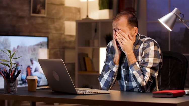 A man rubs his eyes in front of a laptop, feeling tired and sleepy