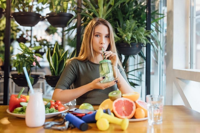 A white blonde woman drinking infused water with fruit slices, plates of fruits and vegetables in front of her
