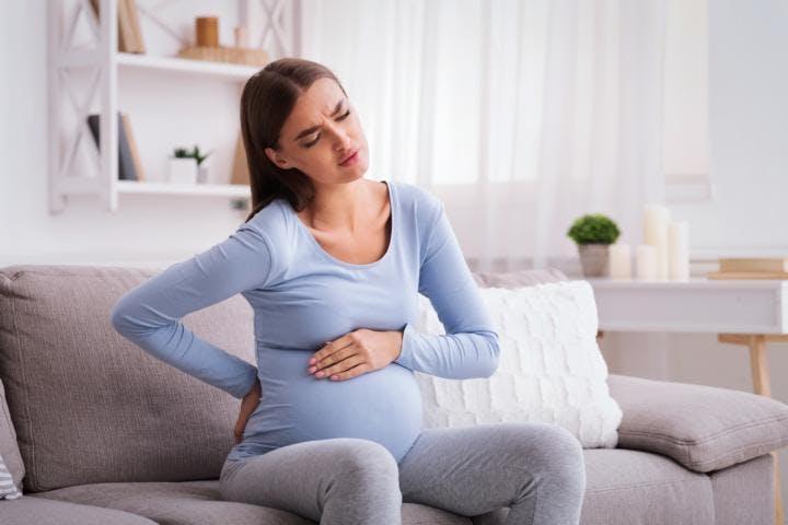 Pregnant woman in pain sitting on a couch with one hand on her belly and one on her back