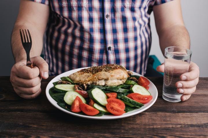 A man eating chicken breast with a side of salad consisting of cucumber and tomatoes