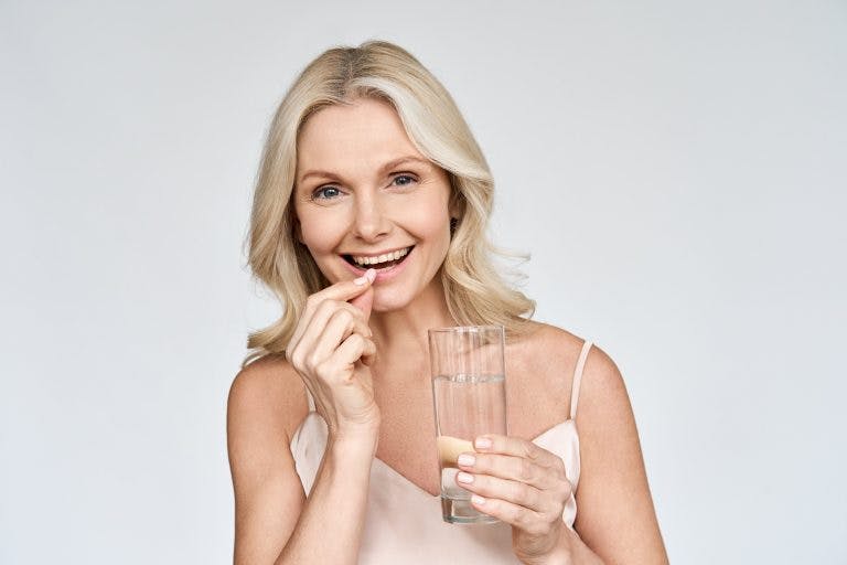 Happy middle aged 50s woman holding pill and glass of water taking dietary supplements