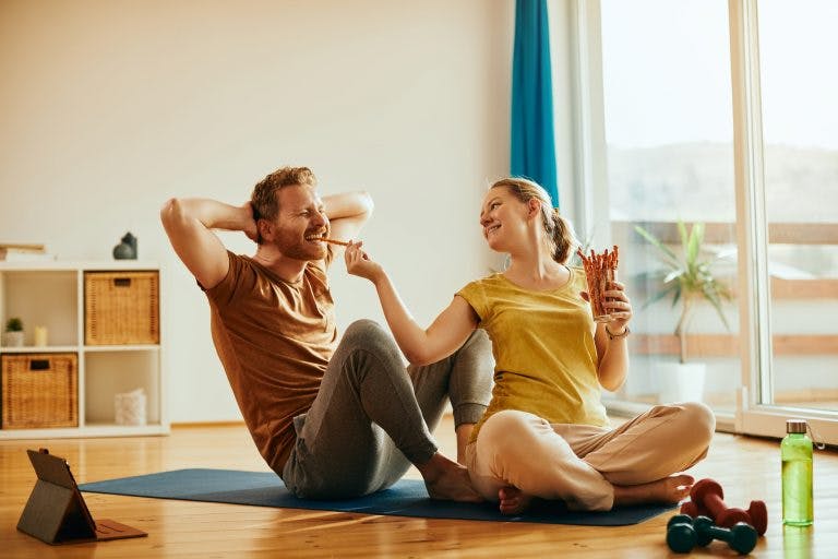 A woman feeding a man snacks while doing exercises at home