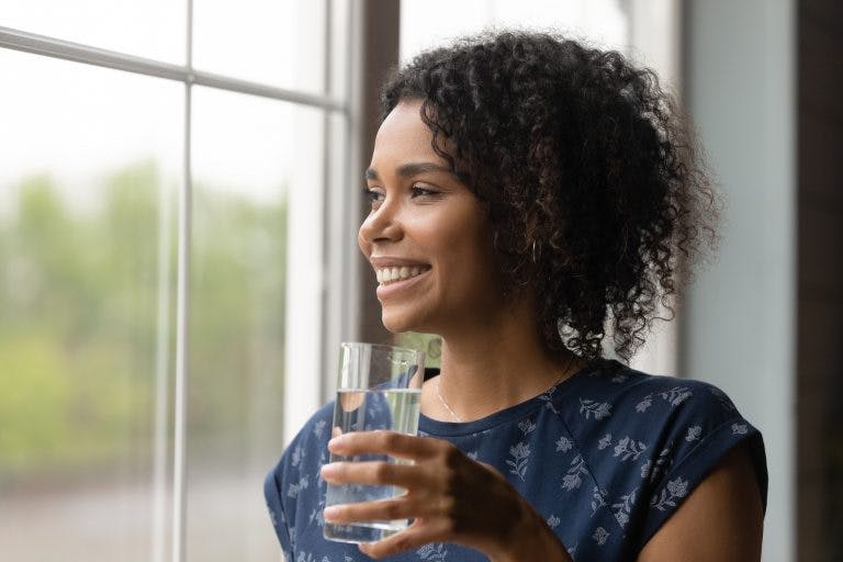 Smiling millennial biracial woman look in distance hold glass of water drink clean clear mineral aqua for body refreshment